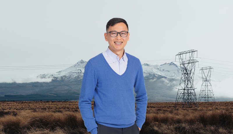 Man in a blue sweater and blue shirt with short black hair standing in front of a landscape shot of a snowy mountain and towers in the background.