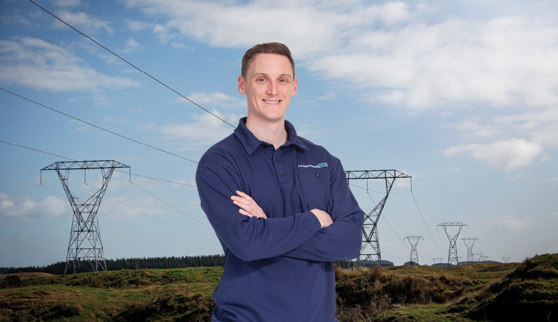 Man in a blue collared shirt with short brown hair standing in front of a landscape shot of grass and a big blue sky with towers and power lines running across it.