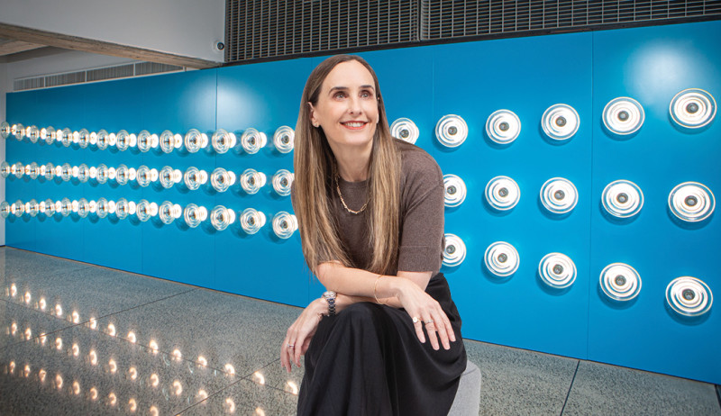 Image of a woman with long brown hair in a brown shirt and black skirt sitting on a stool in front of a blue wall with little lights across it.