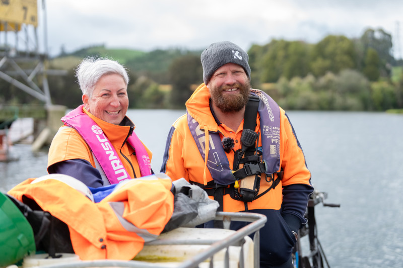 Image of a man and a woman in high-vis standing in front of a transmission tower on a lake.