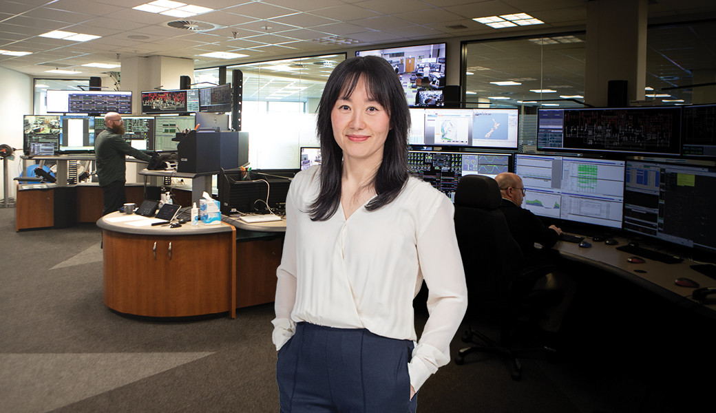 Image of a woman in a white shirt with black hair standing in front of a room filled with desks and computers.