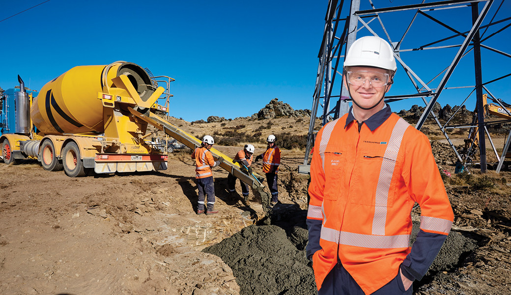 Man in personal protective gear wearing a hard-hat and safety glasses standing in front of a construction site with a concrete truck and tower in the background.