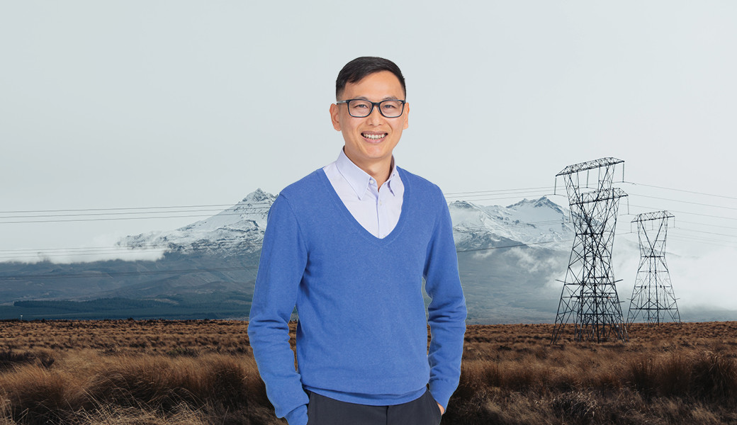 Man with short black hair wearing a blue sweater and blue button up shirt standing in front of a landscape with snowy mountains and power lines in the background
