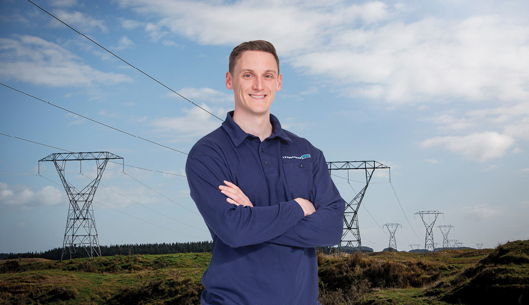 Man in a blue collared shirt with short brown hair standing in front of a landscape of green grass and a big blue sky with power lines and towers running across it.