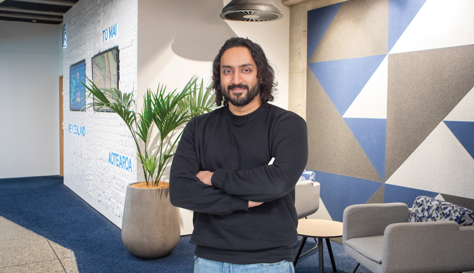 Image of a man with dark hair and beard wearing a black shirt and standing in front of a reception area with his arms crossed.