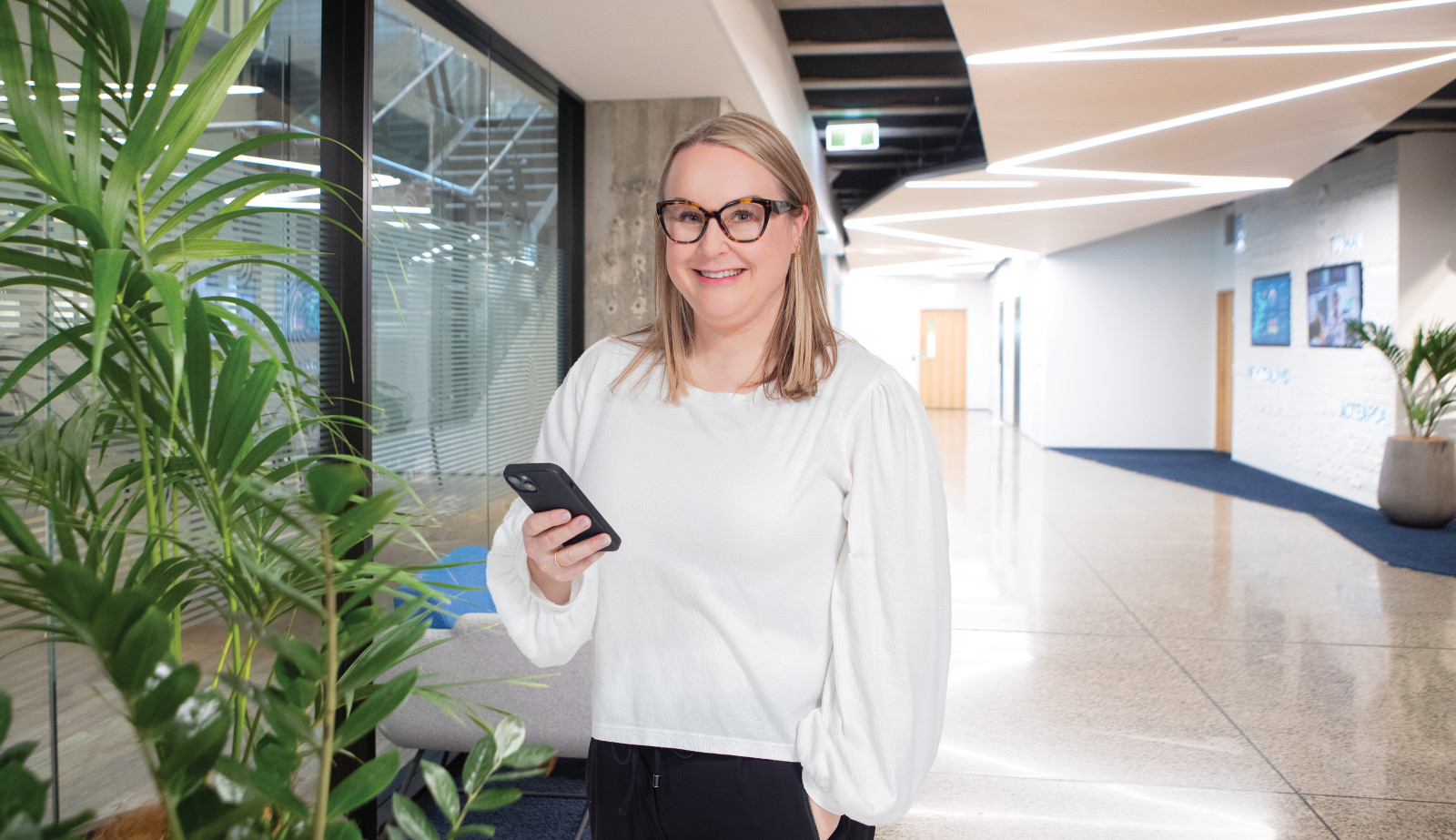 Image of a blonde-haired woman wearing glasses and a white shirt holding a phone while standing in front of a corporate hallway with a large plant at the forefront.