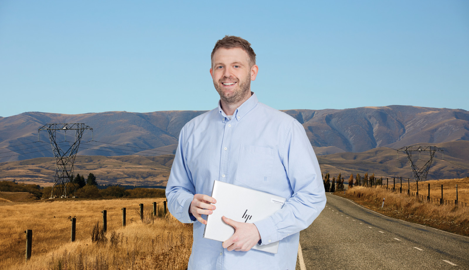 Image of a man with blonde hair wearing a blue button up shirt holding a laptop in front of a landscape shot of dry grass and road and powerlines.