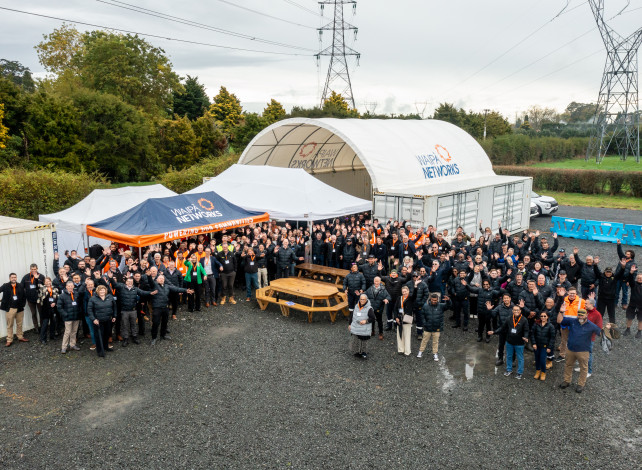 Image of a group of people gathered in a outdoor space near a white tent with powerlines in the background.